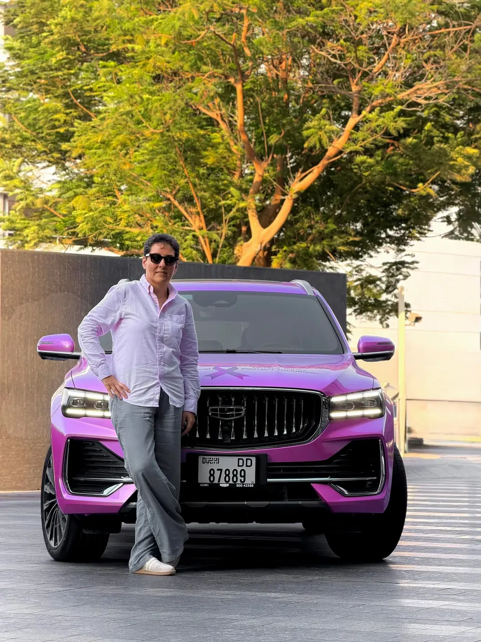 Mehrnoosh in sunglasses leaning on a pink-wrapped Geely SUV carrying a breast cancer awareness ribbon