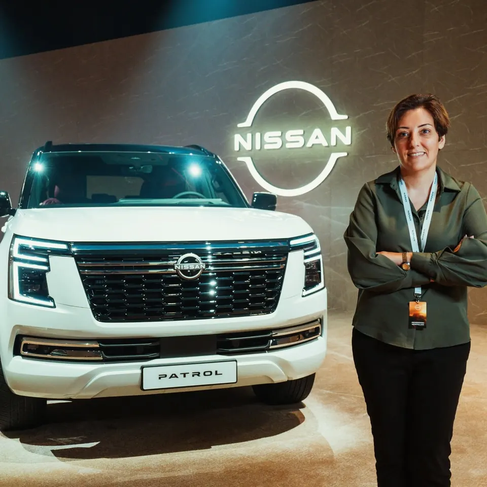 Mehrnoosh with an event lanyard, arms crossed, beside a white Nissan Patrol and an illuminated Nissan logo