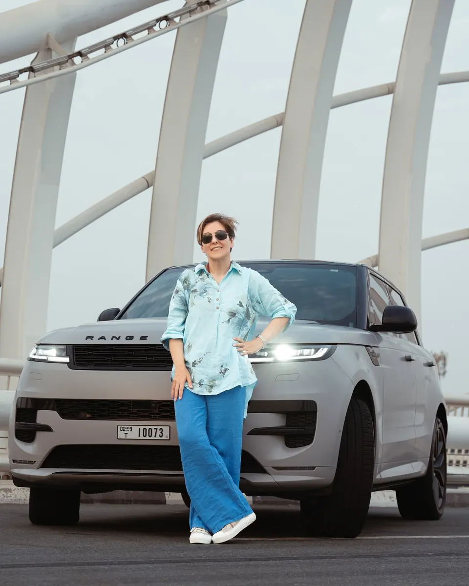 Mehrnoosh leaning on the front of a satin grey Range Rover Sport with its headlights on, under white bridge arches
