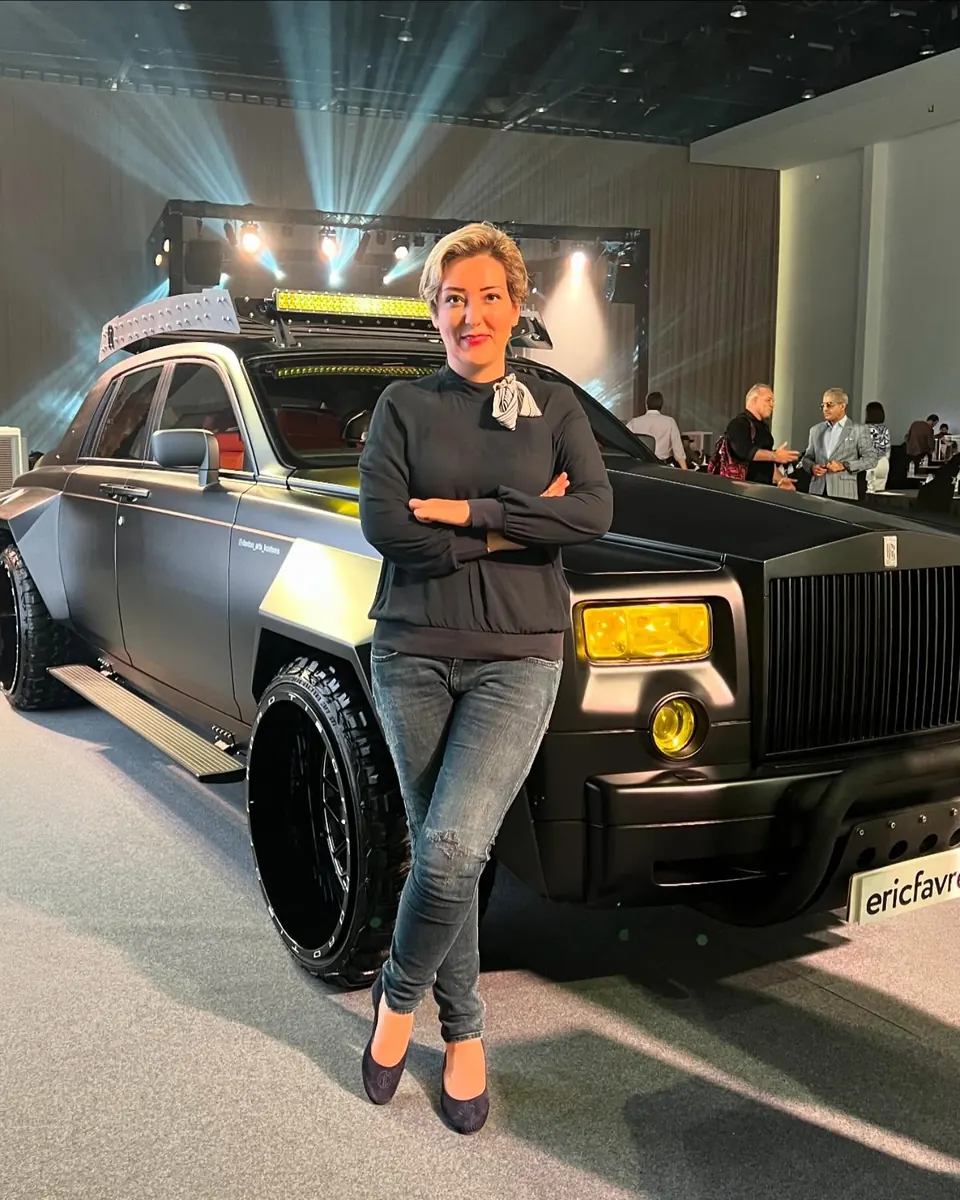 Mehrnoosh, arms crossed, leaning on a matte black custom off-road Rolls-Royce under stage lighting at an automotive show