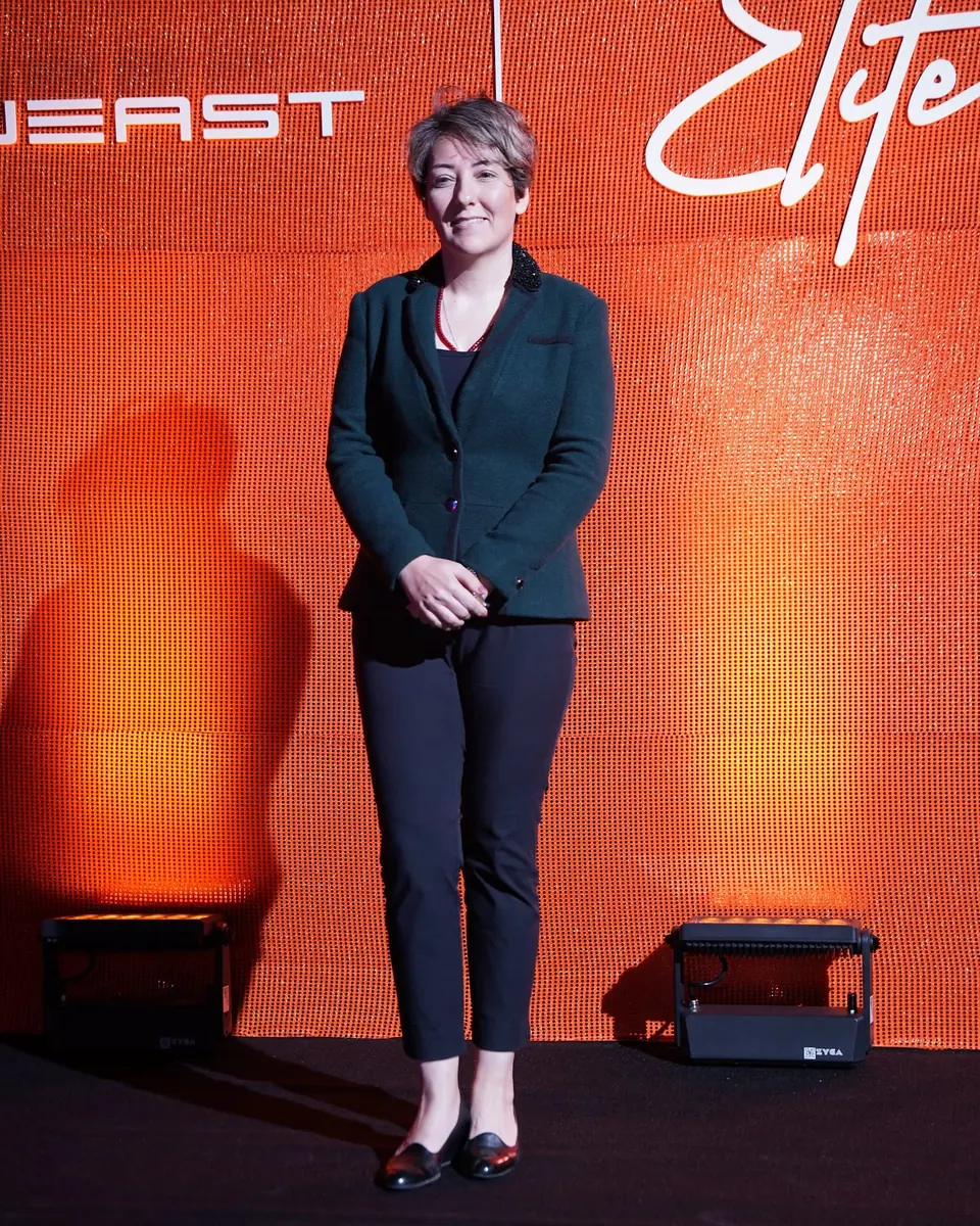 Mehrnoosh in a dark green blazer on an orange sequin stage backdrop with Soueast and Elite branding